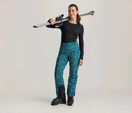 Femme souriante debout avec des skis sur l'épaule, portant un haut noir à manches longues, un pantalon de ski turquoise à motifs et des chaussures de ski noires.