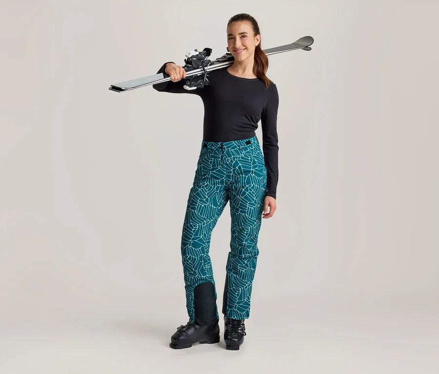 Femme souriante debout avec des skis sur l'épaule, portant un haut noir à manches longues, un pantalon de ski turquoise à motifs et des chaussures de ski noires.