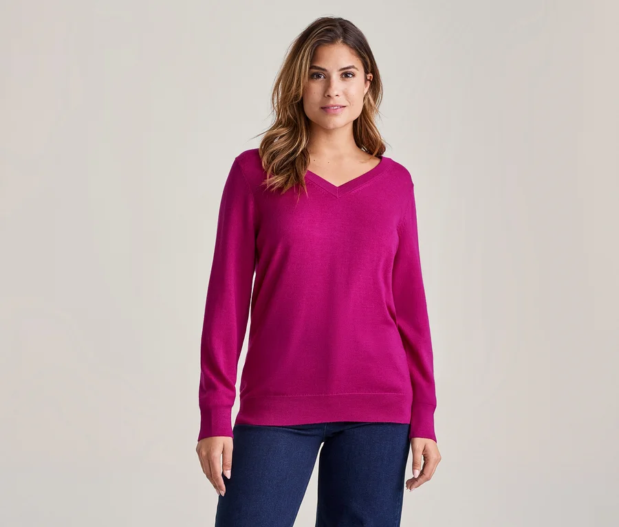 Femme aux cheveux mi-longs portant un pull fuchsia à col en V et un jean bleu foncé devant un fond clair.