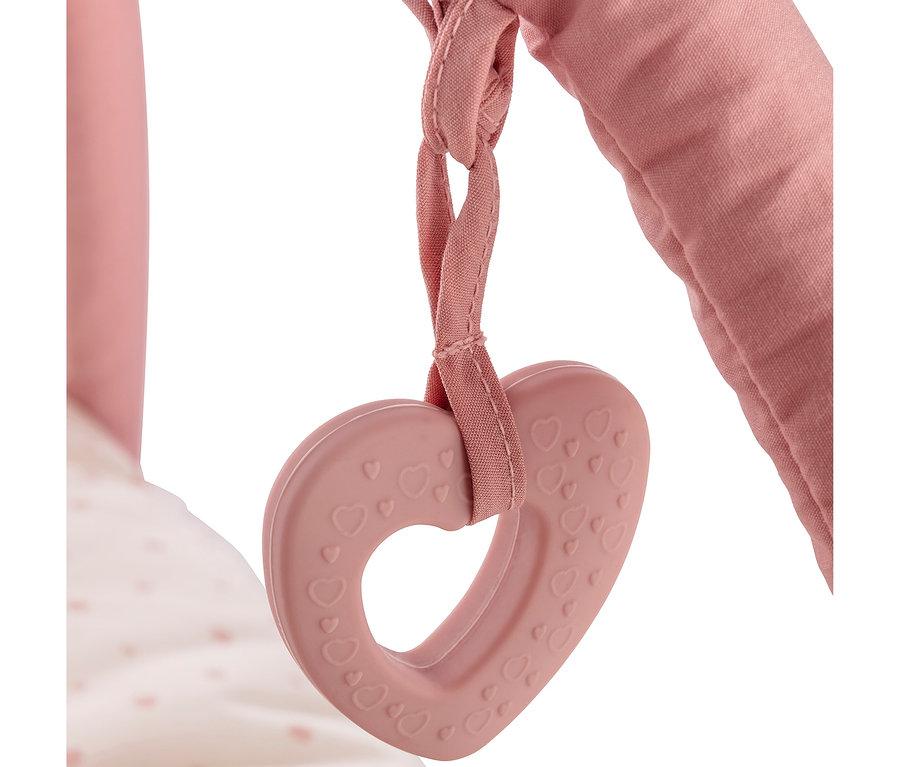 Un anneau de dentition rose en forme de cœur avec de petits cœurs en relief est suspendu à une sangle en tissu rose, attachée à une barre rose rembourrée. Un tissu blanc à pois roses est visible en arrière-plan.
