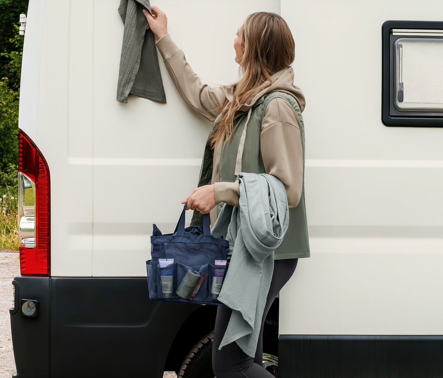 Femme accrochant une serviette en mousseline gris-vert à un camping-car, tenant un sac de douche bleu foncé et une autre serviette en mousseline gris-vert clair.