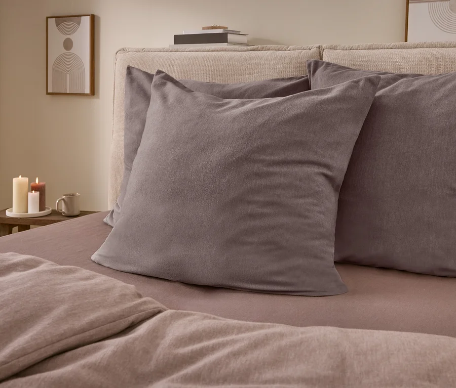 Un lit avec une tête de lit rembourrée claire, deux oreillers gris foncé et une literie marron clair. À côté, une table en bois avec trois bougies allumées, une tasse et un tableau encadré au mur.