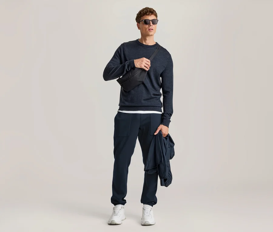 Homme portant un pull bleu foncé, un pantalon bleu foncé, des baskets blanches et des lunettes de soleil, avec une banane noire sur la poitrine et tenant une veste bleu foncé.