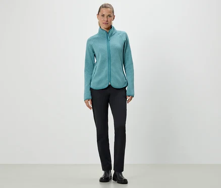 Une femme debout portant une veste en polaire turquoise et un pantalon noir.