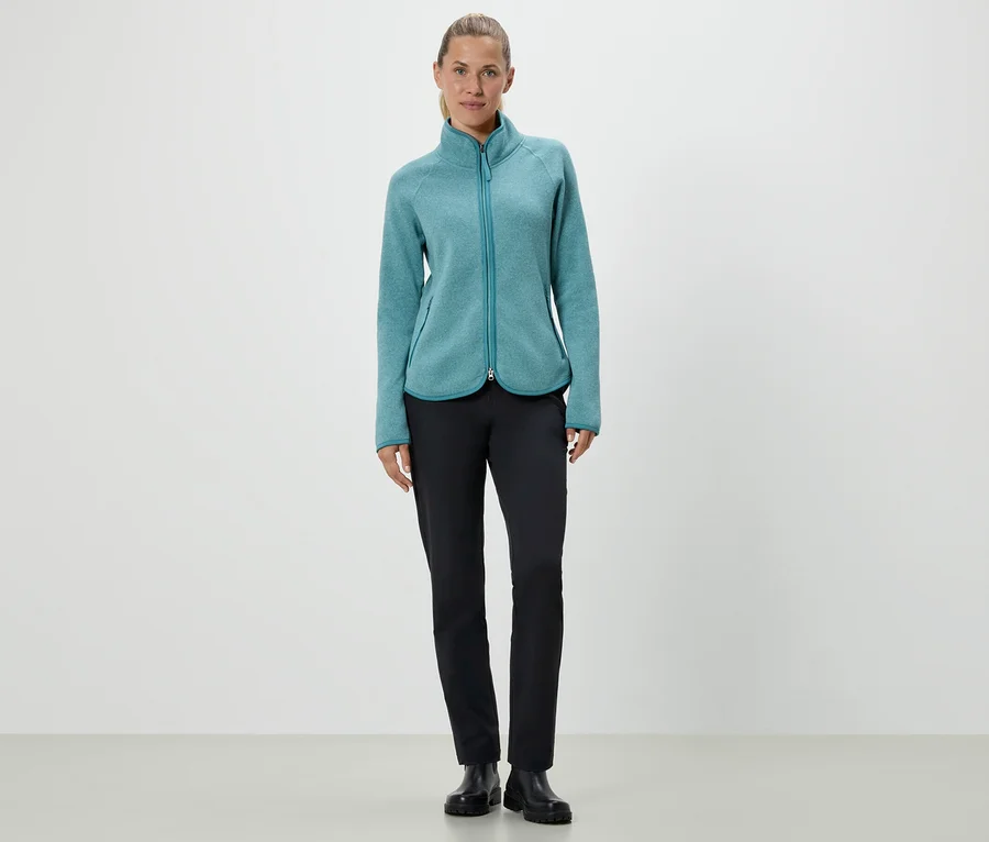 Une femme debout portant une veste en polaire turquoise et un pantalon noir.