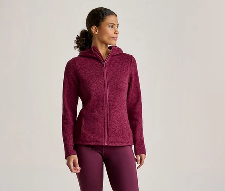 Une femme porte une veste à capuche zippée bordeaux et un legging assorti, debout et regardant vers la droite.