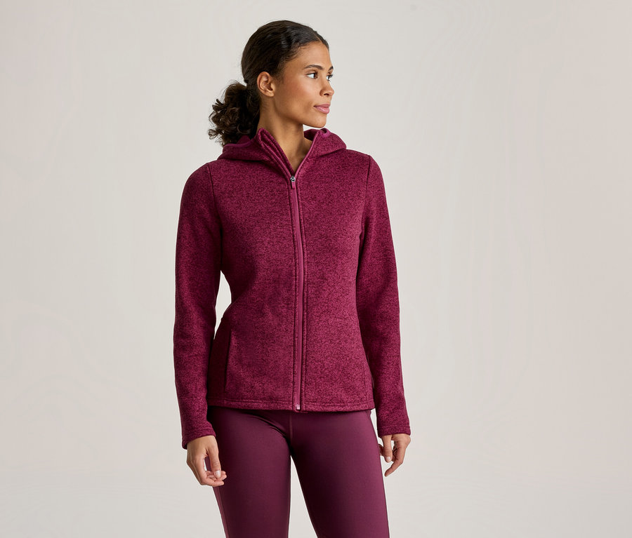 Une femme porte une veste à capuche zippée bordeaux et un legging assorti, debout et regardant vers la droite.