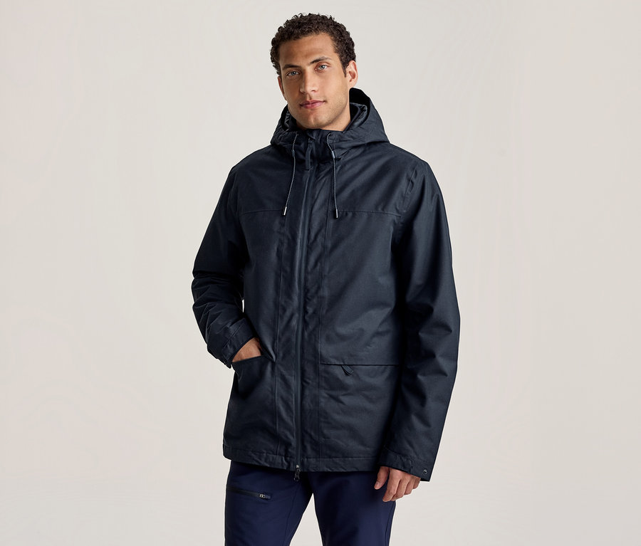 Un homme portant une veste à capuche bleu foncé et un pantalon bleu foncé se tient avec une main dans la poche de sa veste, regardant devant lui.