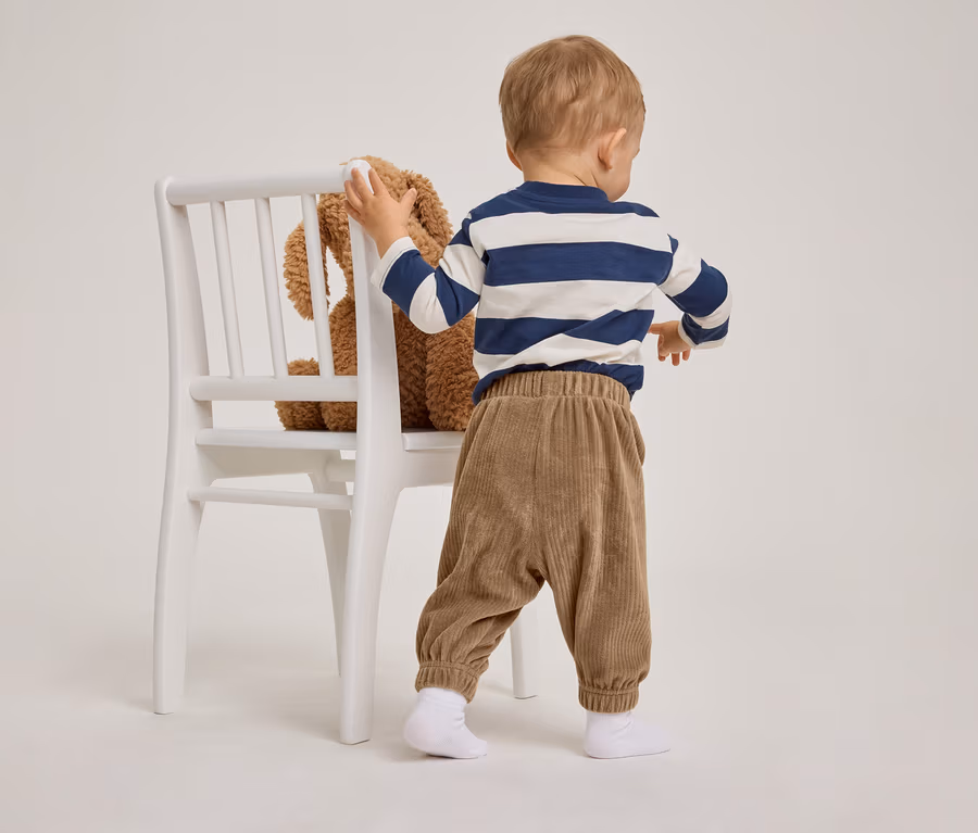 Un tout-petit vu de dos, se tenant au dossier d'une chaise blanche. Il porte un t-shirt à manches longues rayé bleu et blanc et un pantalon en velours côtelé marron. Un ours en peluche marron est assis sur la chaise.