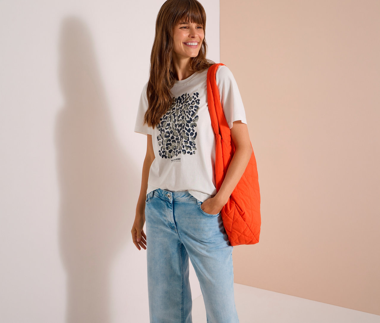 Femme souriante aux longs cheveux bruns portant un T-shirt en jersey CECIL blanc à imprimé léopard, un jean bleu clair et un sac fourre-tout matelassé orange sur l'épaule.