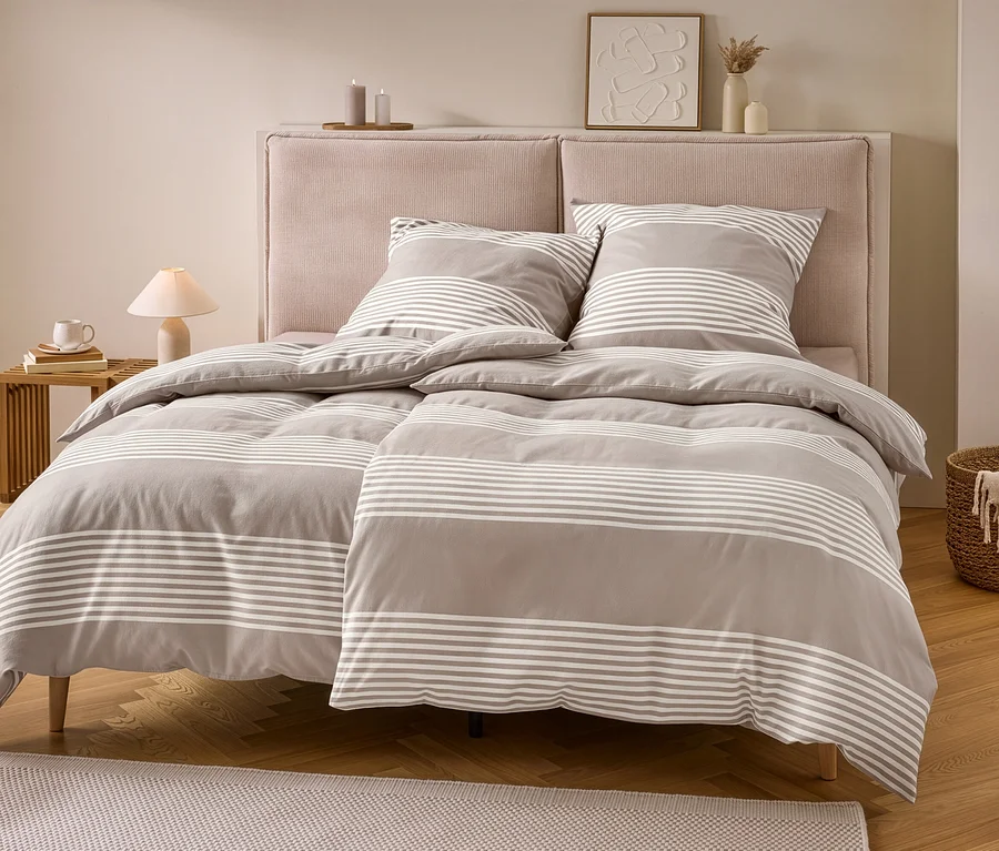 Un lit avec une parure de lit en molleton grise à rayures blanches, comprenant deux oreillers et deux couettes. Le lit a une tête de lit rose, une table de chevet avec une lampe et un panier avec une couverture.