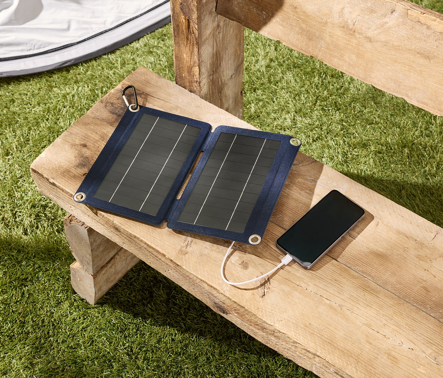 Un chargeur de panneau solaire recharge un smartphone posé sur un banc en bois à l'extérieur.