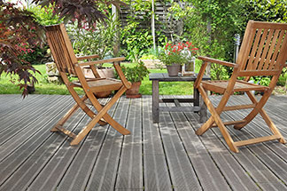 Deux chaises de jardin en bois se trouvent sur une terrasse donnant sur un jardin verdoyant – devant elles, une petite table carrée en bois sur laquelle se trouve un vase de fleurs.