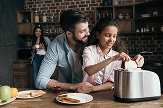 Petit-déjeuner ensemble : un jeune père met deux tranches de pain grillé dans un grille-pain couleur argent posé sur le comptoir de la cuisine.