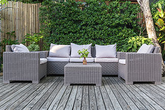 Meubles de salon pour le jardin : sur une terrasse en bois se trouvent un canapé gris, une table et deux fauteuils décorés de coussins blancs.