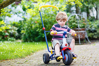 Utilisez les jouets d'extérieur à l'âge approprié : un petit garçon portant un short et une chemise à rayures roses et bleues conduit son tricycle le long d'un chemin pavé dans le jardin.