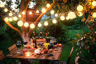 L'éclairage de jardin comme décoration : une table de jardin en bois placée sous un arbre et recouverte de vin, de fromage et de fruits. Au-dessus de celle-ci est suspendue une guirlande lumineuse allumée avec des lampions ronds.