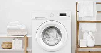 Lave-linge avec du linge de bain dans le tambour, à côté se trouvent des étagères en bois avec de la lessive et des serviettes.