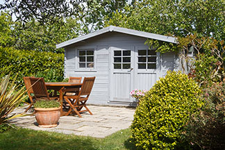 Rangez les meubles de salon dans l'abri de jardin : dans un jardin aux hautes haies vertes se trouve une maison d'été grise, devant laquelle se trouve une table ronde en bois avec quatre chaises en bois.