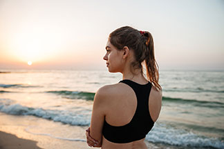 Vue arrière d'une jeune femme en soutien-gorge de sport noir, debout au bord de la mer, contemplant le coucher du soleil.