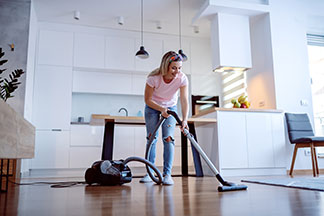 Une femme portant un short en denim et un t-shirt rose nettoie le sol en bois de la salle à manger avec un aspirateur – la cuisine blanche est visible en arrière-plan.