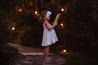 Éclairage de jardin de bonne qualité : une petite fille en robe d'été est debout dans le jardin, éclairée par une guirlande de lampions et lit un livre.