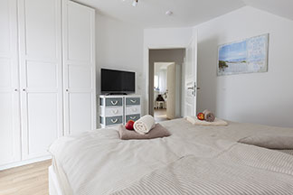 Un design intemporel : dans une chambre à coucher meublée de façon lumineuse, il y a une armoire blanche à trois portes et une commode avec des tiroirs gris et blancs sur laquelle est placée une télévision.