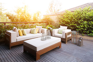 Devant une porte de terrasse ouverte il y a des meubles de salon en rotin synthétique tissé et deux coussins d’assise. La table est dressée et plusieurs coussins décoratifs sont disposés sur le canapé.
