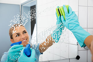 Nettoyez correctement le miroirs de salles de bain : une jeune femme nettoie son miroir avec des gants bleus et un spray nettoyant.