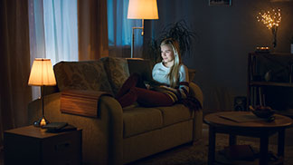 Bonne qualité pour l'éclairage de votre salon : une jeune femme blonde est allongée sur son canapé et regarde son ordinateur portable tandis que le salon est faiblement éclairé par trois lampes.