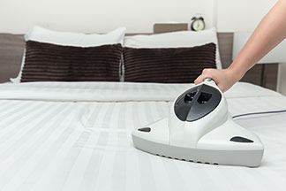 La main d'une femme guide un aspirateur à main blanc sur un lit recouvert de draps blancs.