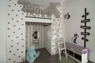 Espace de rangement et surface d’assise : un lowboard blanc contre le mur dans la chambre des enfants, a des tiroirs avec des compartiments en bas et un coussin d'assise violet.