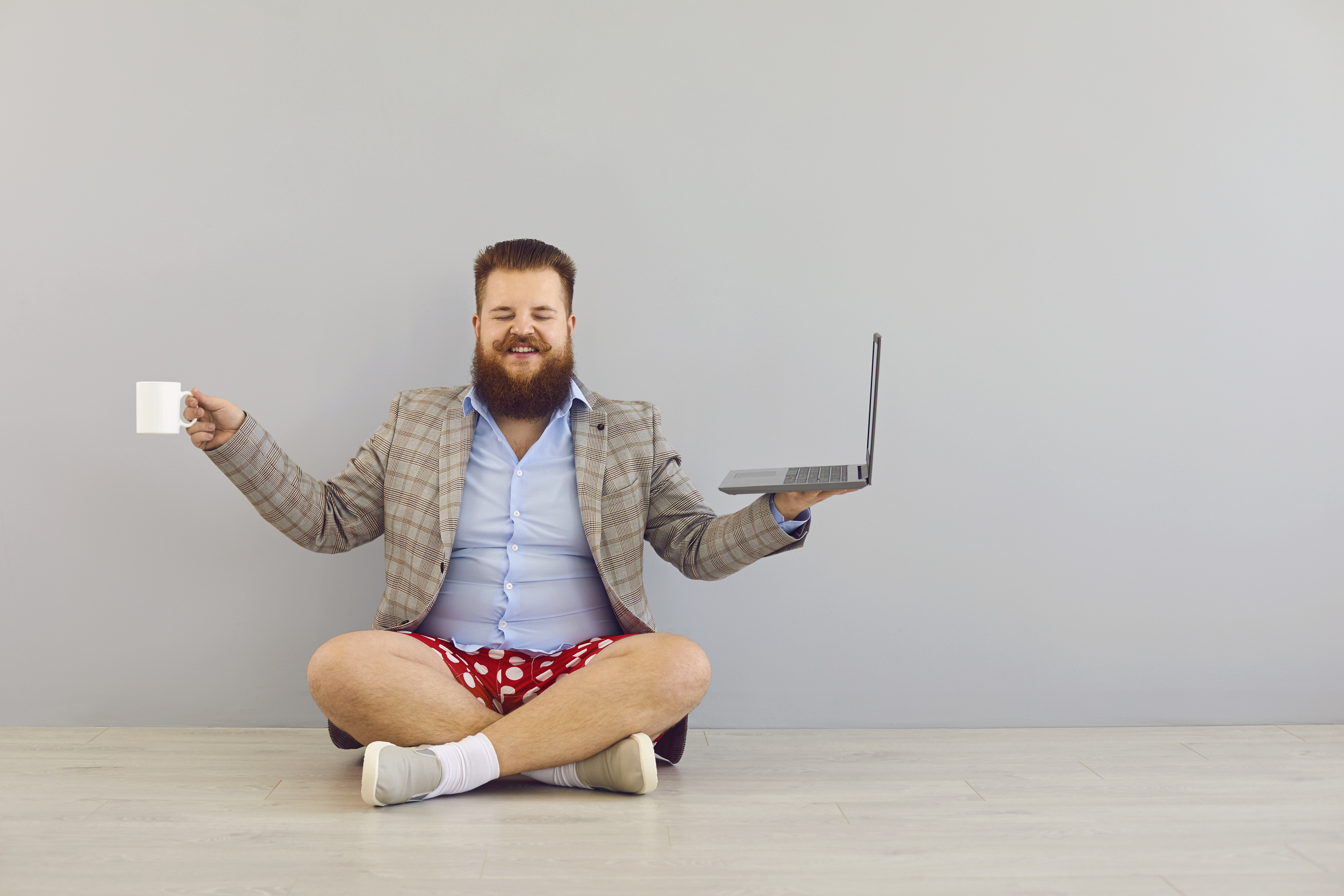 Homme assis en tailleur avec un ordinateur portable, une tasse de café et portant un veston, une chemise et un boxer.