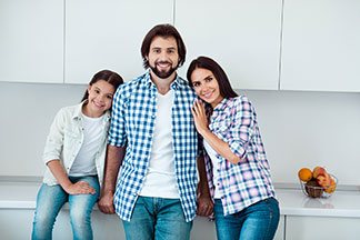 Trouver la bonne cuisine : un jeune homme, une femme et leur jeune fille se tiennent debout en riant, le dos tourné au plan de travail de leur cuisine blanche équipée.