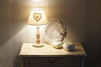Une table de chevet rectangulaire blanche avec un tiroir, sur laquelle se trouvent deux cœurs en céramique, une couronne en bois tressé et une lampe, se dresse dans le couloir devant un mur blanc.