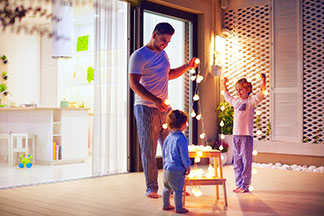 Au crépuscule, un père, son jeune fils et sa jeune fille sont debout sur la terrasse, s'occupent d'une guirlande lumineuse avec de petites lampes rondes.