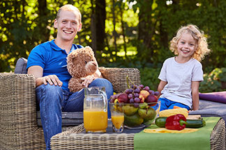 Un père est assis avec sa jeune fille sur des meubles de jardin en rotin – devant eux se trouve une table en rotin avec un bol de fruits, des légumes et du jus d'orange.