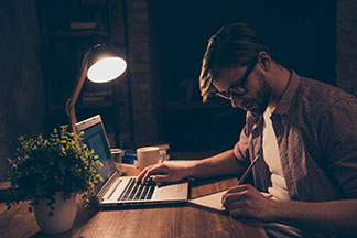 Lampe de table sur le bureau : un jeune homme est assis à un bureau en bois bien éclairé avec un stylo et un bloc-notes devant un ordinateur portable – il fait sombre autour de lui.