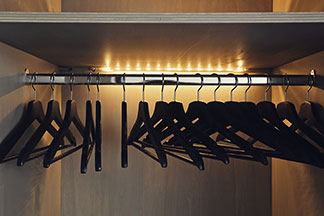 Lampes d'armoire chez Tchibo dans une grande qualité : une tringle à vêtements dans une armoire en bois sombre avec des cintres vides – éclairée par une bande de lumière sur le mur du fond.