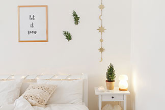 Lampes de qualité Tchibo: une chambre à coucher meublée en blanc avec un lit et une table de chevet sur laquelle se trouve une lampe décorative allumée en forme de champignon.