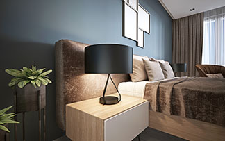 Une lumière bien-être sur la table de chevet : une table de chevet rectangulaire en bois avec une lampe ronde se trouve à côté d'un lit double sur lequel reposent un couvre-lit marron et des oreillers décoratifs beiges.