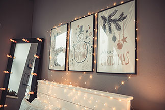 Des lampes décoratives pour les photos et tableaux : sur un mur gris et au-dessus d'un lit blanc sont accrochés trois tableaux dans des cadres noirs, entourés d'une série de lumières.