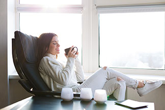 Se détendre dans le fauteuil : une femme vêtue de blanc est assise devant une fenêtre, dans un fauteuil de relaxation en cuir noir, et tient une tasse de thé.