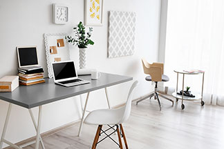 Le bon espace de travail : un bureau gris sur lequel se trouve un ordinateur portable est placé contre un mur blanc sur lequel sont accrochés deux tableaux décoratifs et une horloge – devant, une chaise blanche.