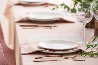 Une table décorée de façon festive, avec une nappe de lin rose. La table est dressée avec des assiettes plates et des couverts en cuivre.