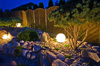 Utilisez correctement l'éclairage du jardin : des lampes de jardin rondes se dressent contre une clôture en bois, éclairant de petits arbres d'ornement.