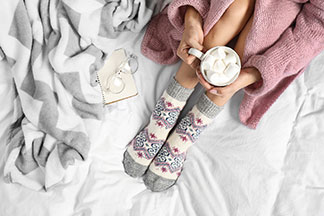 Pour la saison fraîche : une femme en peignoir rose, aux collants épais à motifs et aux jambes serrées, est assise avec une tasse blanche pleine de guimauve sur son drap de lit.