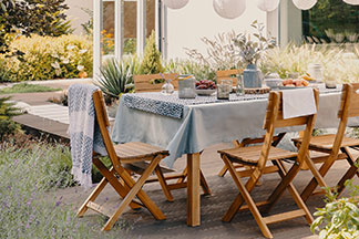Un espace repas accueillant en plein air : sur une terrasse, il y a une table dressée avec une nappe bleue et cinq chaises de jardin en bois.