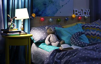 Une jeune mère est allongée dans son lit avec son enfant en train de lire un livre pendant que la lampe de chevet les éclaire.