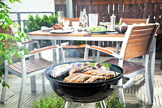 Un balcon sur lequel se trouve un grill avec de la viande. En arrière-plan il y a une table de jardin rustique avec des chaises en bois.